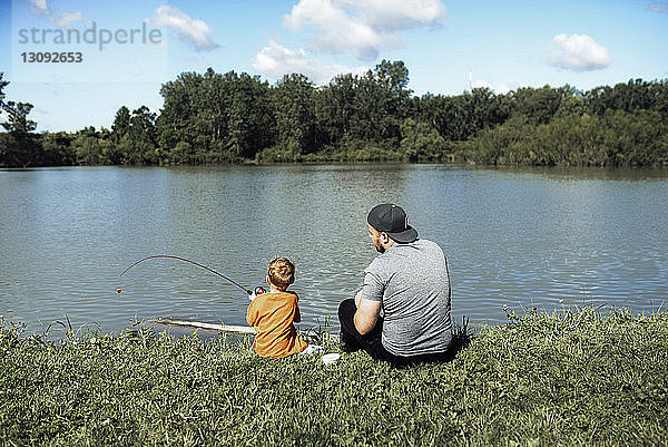 Rückansicht des Sohnes beim Angeln  während er mit dem Vater am Seeufer gegen den Himmel sitzt