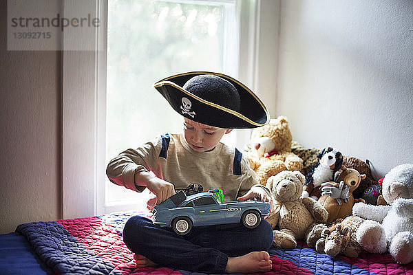 Junge mit Piratenhut spielt mit Spielzeugauto  während er zu Hause auf dem Bett sitzt