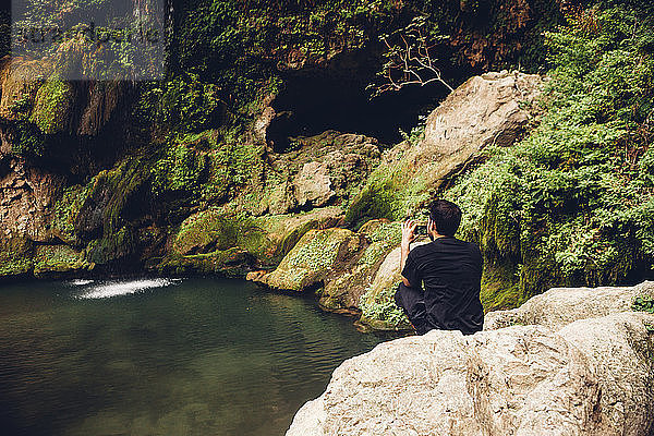 Rückansicht eines männlichen Wanderers  der mit seinem Mobiltelefon fotografiert  während er auf Felsen am Flussufer sitzt