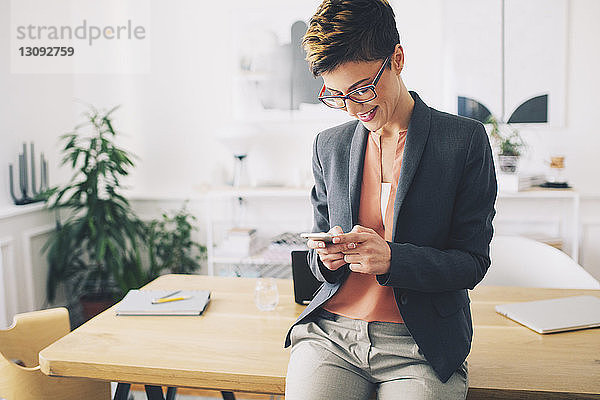 Glückliche Geschäftsfrau benutzt Mobiltelefon  während sie im Büro am Tisch steht