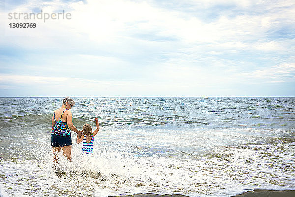 Rückansicht der Enkelin und der Großmutter  die sich beim Strandspaziergang am Ufer an den Händen halten