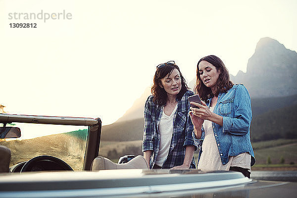 Frau zeigt Freundin Mobiltelefon im Cabriolet bei Sonnenuntergang