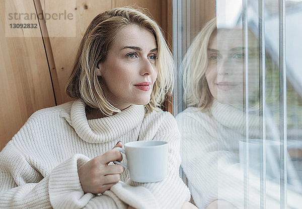 Frau hält Kaffeetasse  während sie zu Hause durchs Fenster schaut