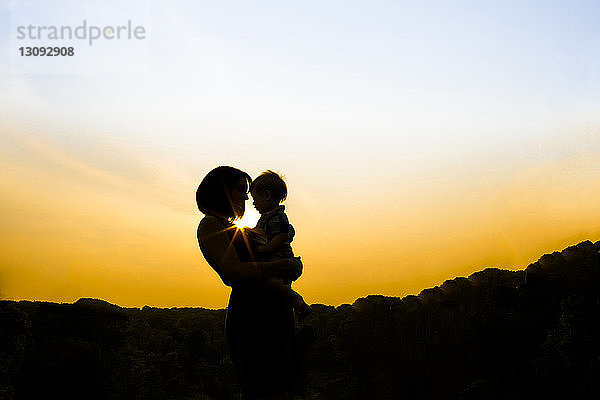 Silhouette einer Mutter  die einen Sohn trägt  während sie bei Sonnenuntergang im Park gegen den Himmel steht