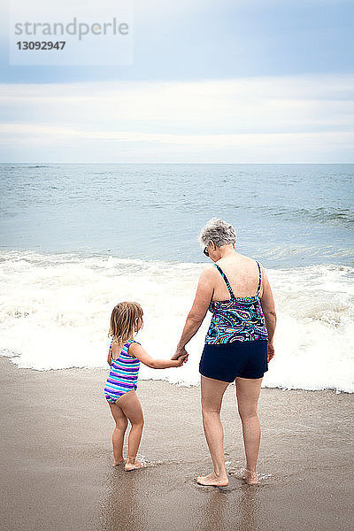 Enkelin und Großmutter halten Händchen  während sie am Strand am Ufer stehen
