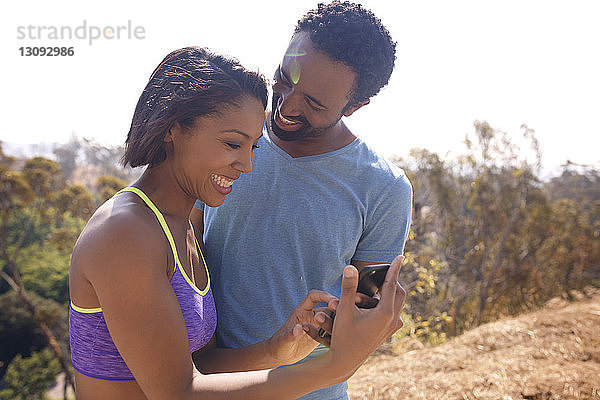 Glückliches Paar beim SMS-Versand im Stehen auf dem Feld vor klarem Himmel