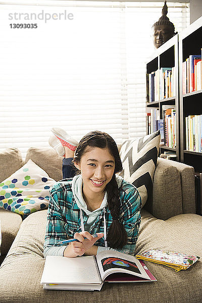 Porträt einer selbstbewussten Teenagerin  die Hausaufgaben macht  während sie zu Hause auf der Couch liegt