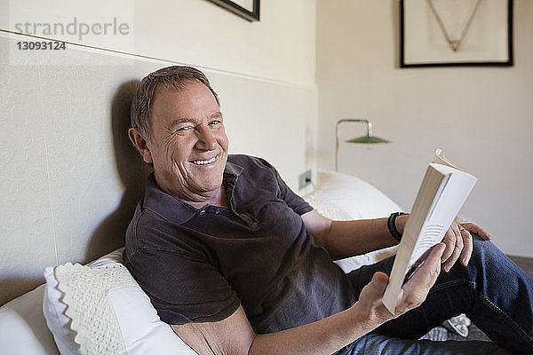Porträt eines glücklichen älteren Mannes  der zu Hause auf dem Bett sitzend ein Buch in der Hand hält