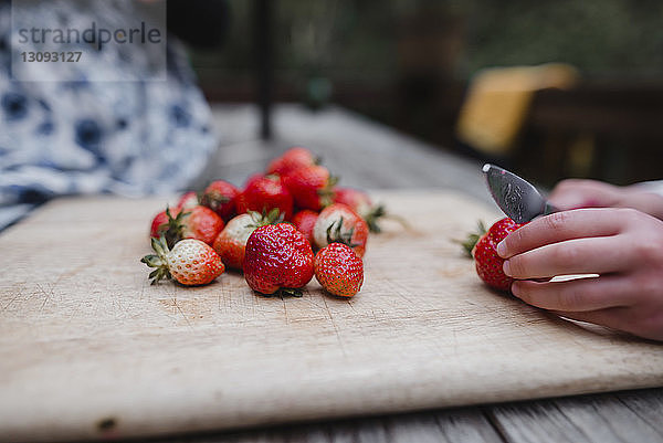 Geschnittene Hände eines Mädchens  das Erdbeeren auf einem Schneidebrett bei Tisch schneidet