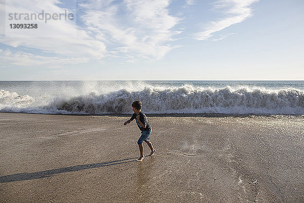 Junge in voller Länge rennt vor den rauschenden Wellen am Strand gegen den Himmel