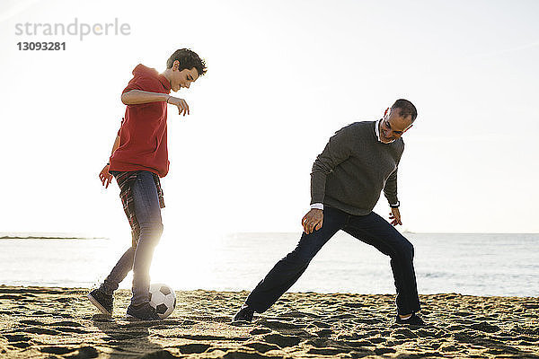 Vater und Sohn spielen bei strahlendem Sonnenschein am Strand bei klarem Himmel Fussball