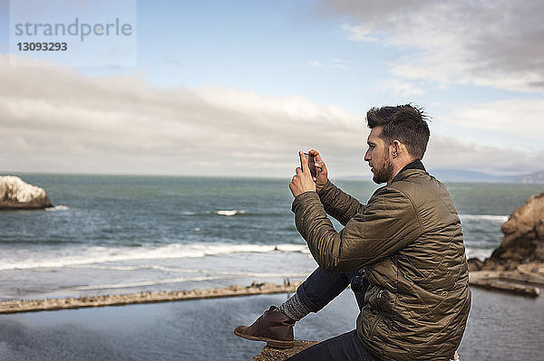 Seitenansicht eines Mannes  der das Meer mit einem Smartphone gegen den Himmel fotografiert