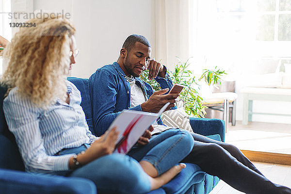 Frau liest Zeitschrift  während der Mann zu Hause auf dem Sofa ein Smartphone benutzt