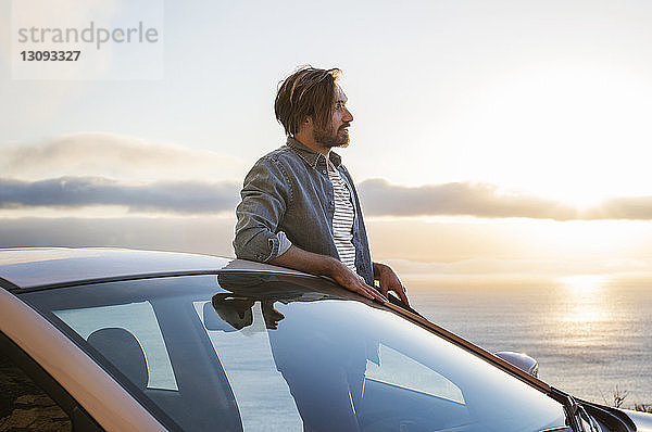 Junger Mann schaut weg  während er bei Sonnenuntergang vor dem Auto am Meer steht