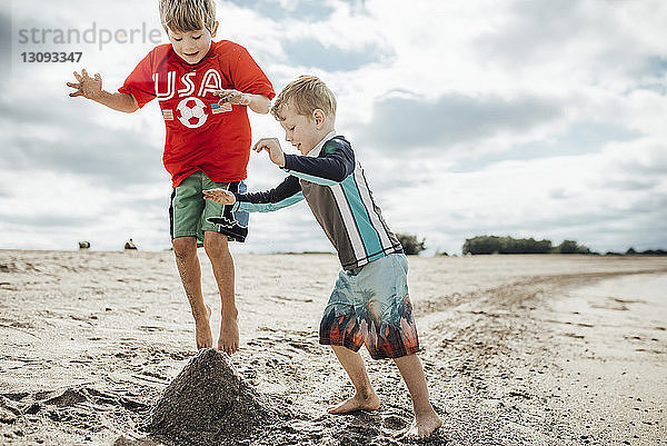 Glückliche Brüder spielen mit Sand am Strand gegen bewölkten Himmel