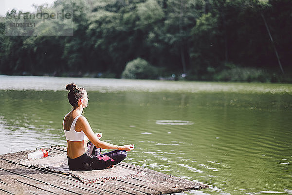 Seitenansicht einer Frau  die meditiert  während sie auf einem Steg über einem See im Wald sitzt