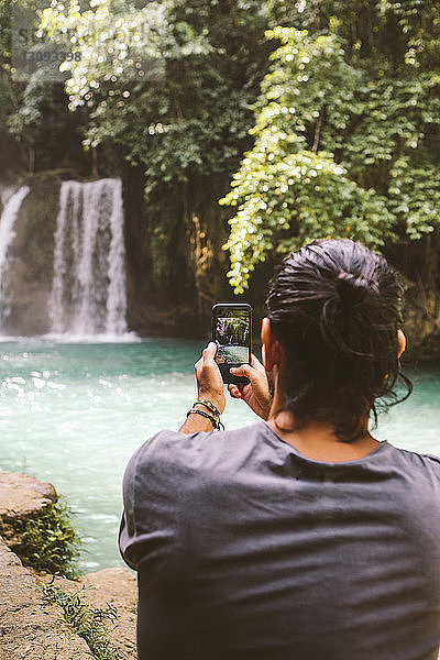 Rückansicht eines Mannes  der im Wald einen Wasserfall mit einem Smartphone fotografiert