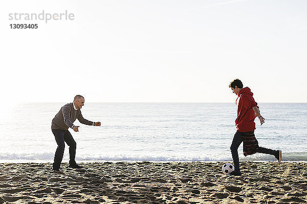 Sohn kickt Fussball  während der Vater am Strand gegen den klaren Himmel verteidigt