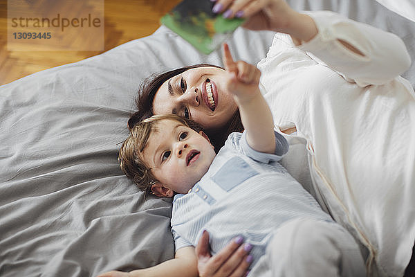 Hochwinkelansicht einer glücklichen Mutter  die mit dem kleinen Jungen liegt und auf ein Bilderbuch auf dem Bett zeigt