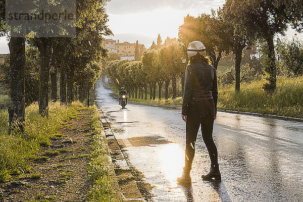 Rückansicht einer Frau  die steht  während ihr Freund bei Sonnenuntergang auf nasser Straße Motorrad fährt