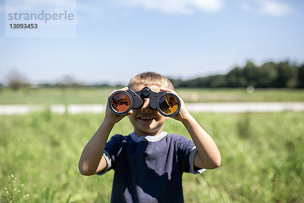 Verspielter Junge  der durch ein Fernglas schaut  während er an einem sonnigen Tag auf dem Feld steht