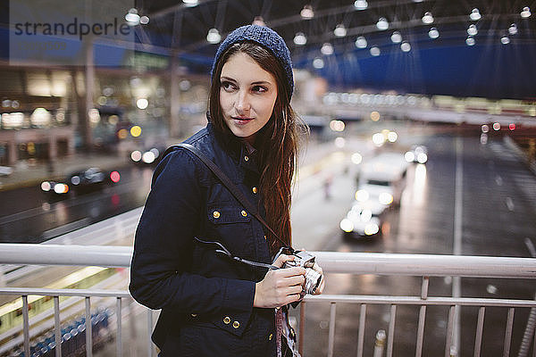 Frau hält Kamera und schaut weg  während sie sich am Geländer im Flughafen anlehnt