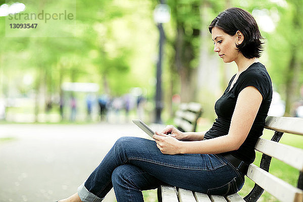 Seitenansicht einer Frau  die einen Tablet-Computer auf einer Parkbank benutzt