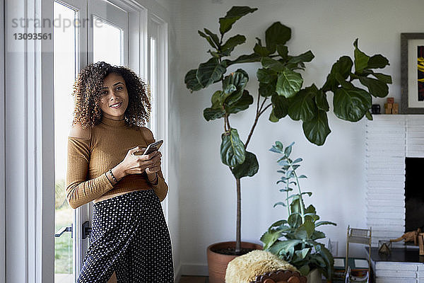 Porträt einer lächelnden Frau  die ein Mobiltelefon benutzt  während sie zu Hause am Fenster steht