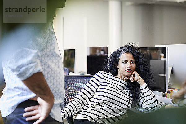 Geschäftsfrau mit Hand am Kinn  die am Schreibtisch sitzend von einem männlichen Kollegen im Büro wegschaut
