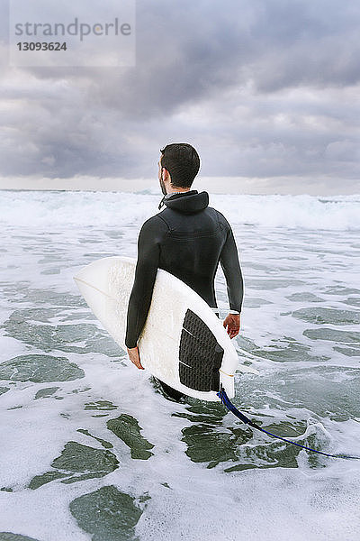 Rückansicht eines Mannes  der ein Surfbrett trägt  während er im Meer gegen einen bewölkten Himmel läuft