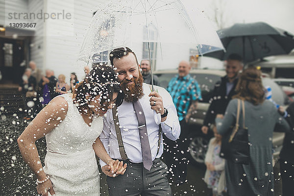 Glückliches frisch verheiratetes Paar geht mit einem Regenschirm spazieren  während die Menschen während der Hochzeitszeremonie im Hintergrund stehen