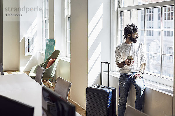 Geschäftsmann schaut durch ein Fenster  während er im Kreativbüro ein Smartphone in der Hand hält