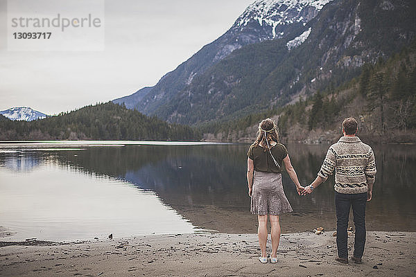 Rückansicht eines jungen Paares  das am Seeufer gegen Berge und Himmel steht und dabei Händchen hält