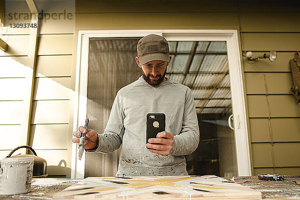 Selbstbewusster Künstler fotografiert Holzkunst mit dem Handy an der Werkbank