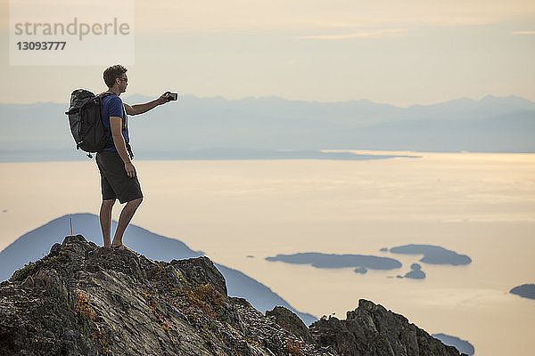 Seitenansicht eines Wanderers mit Rucksack beim Fotografieren  während er auf dem Berg gegen den Himmel steht