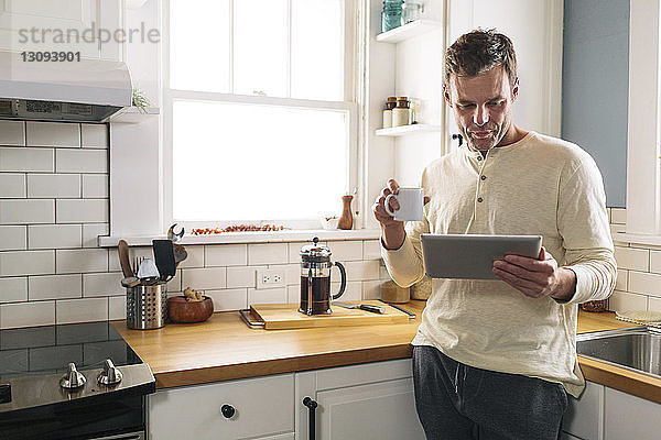 Mann benutzt Tablet-Computer  während er sich zu Hause auf die Küchentheke lehnt