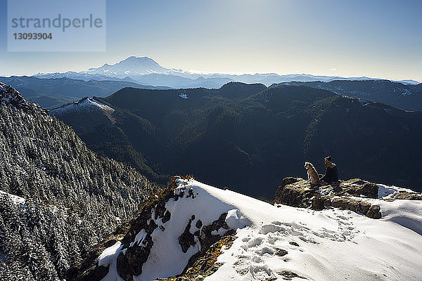 Hochwinkelaufnahme eines Mannes  der im Winter mit Hund auf einem Berg sitzt