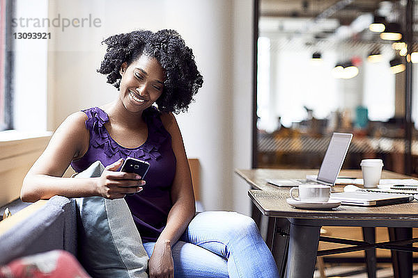 Glückliche Geschäftsfrau schaut auf Smartphone  während sie in der Büro-Cafeteria sitzt