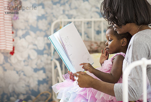 Mädchen zeigt auf ein Bilderbuch  während sie mit der Mutter im Schlafzimmer sitzt