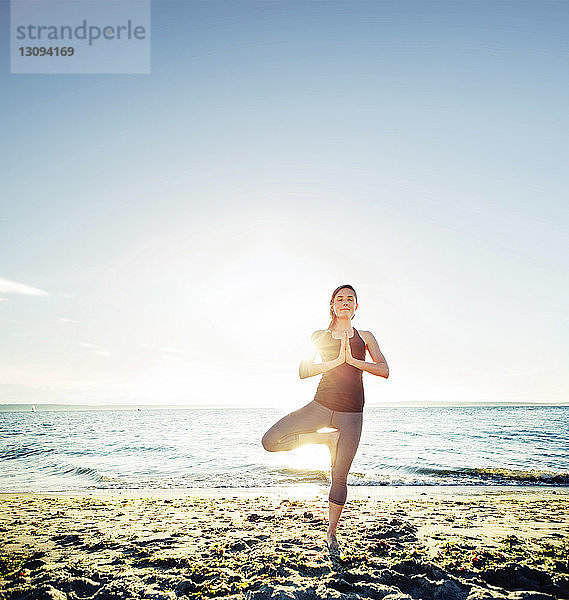 Frau meditiert in Baum-Pose am Strand gegen den Himmel