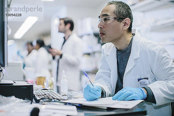 Männlicher Arzt schreibt am Schreibtisch  während seine Kollegen im Hintergrund arbeiten