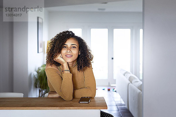 Porträt einer Frau mit Mobiltelefon auf Tisch sitzend zu Hause