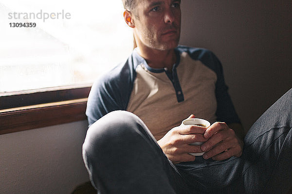 Nachdenklicher Mann mit Kaffeetasse  der zu Hause am Fenster sitzt