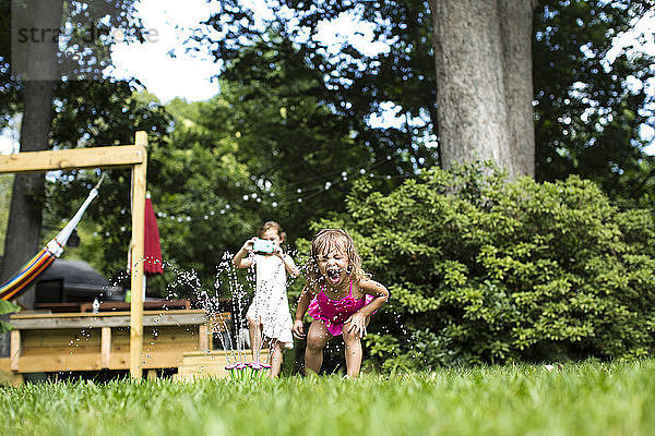 Mädchen fotografiert  während die Schwester im Garten mit der Sprinkleranlage spielt