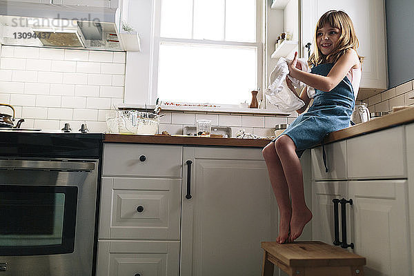 Lächelndes Mädchen reibt sich die Hände mit einer Serviette  während es zu Hause auf dem Küchentisch sitzt