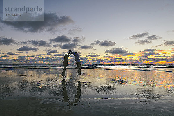 Seitenansicht eines Paares  das in Long Beach bei Sonnenuntergang vor bewölktem Himmel gemeinsam springt