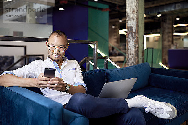 Lächelnder Geschäftsmann benutzt Mobiltelefon  während er im Büro auf dem Sofa sitzt