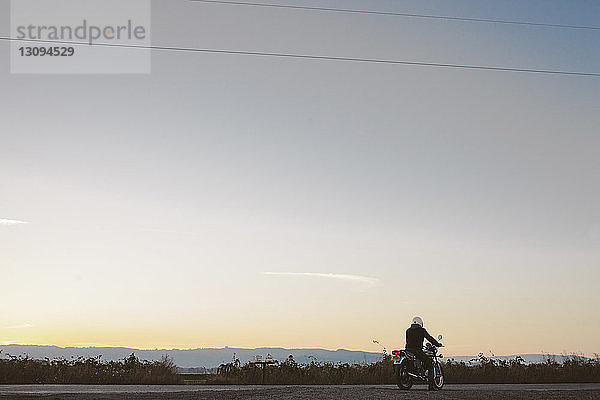 Rückansicht eines männlichen Radfahrers  der bei Sonnenuntergang auf einem Fahrrad auf der Straße gegen den Himmel sitzt