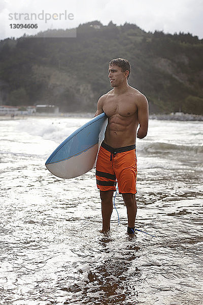 Nachdenklicher behinderter Mann trägt Surfbrett  während er am Ufer steht