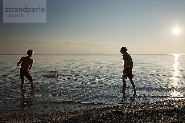 Rückansicht von Jungen  die bei Sonnenuntergang am Strand am Ufer spielen